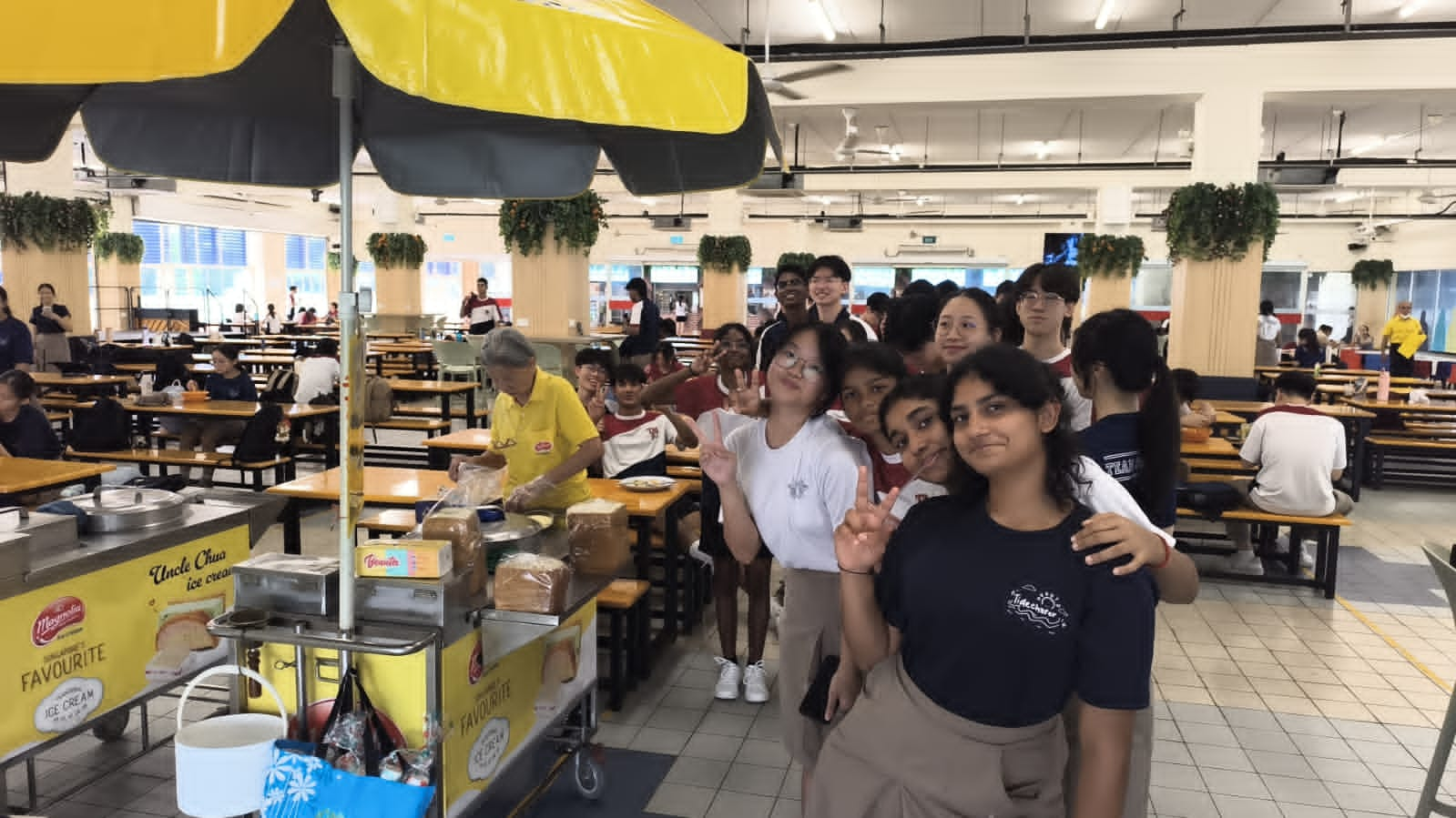 A cheerful group of students queue and pose with peace signs at an "Uncle Chua Ice Cream" cart in a school canteen, with the elderly ice cream vendor serving in the background.